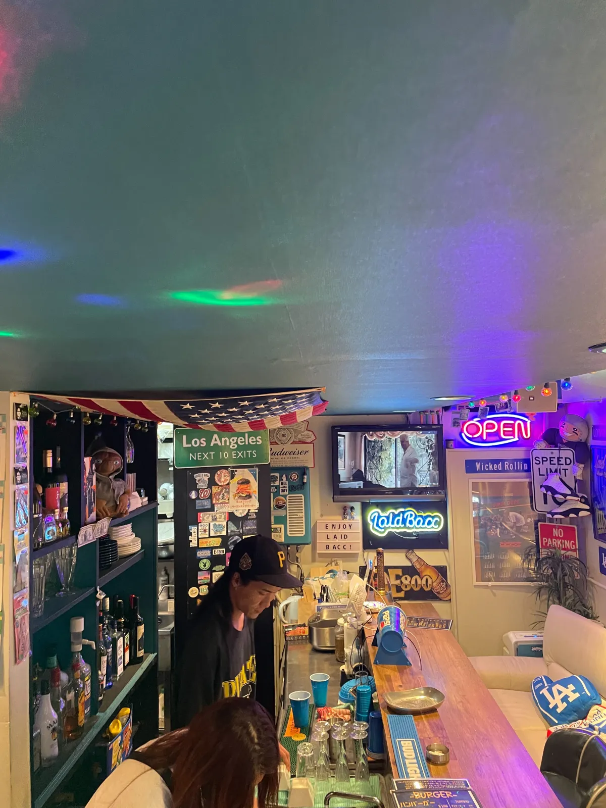 バーテンダーが立つカウンター。Los Angeles Next 10 Exits の標識、OPENネオン、ENJOY LAID BACC のサイン